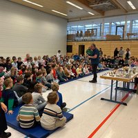 Eine große Gruppe von Kindern sitzt auf Matten in einem Raum und schaut einem Vortragenden zu. Der Vortragende steht an einem Tisch, auf dem verschiedene Materialien oder Modelle liegen. Hinter den Kindern sind weitere Erwachsene zu sehen, die ebenfalls dem Vortrag folgen.