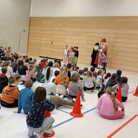 Eine große Gruppe von Kindern sitzt auf dem Boden in einer Sporthalle und schaut auf eine Bühne. Dort stehen zwei Personen, eine in einem roten Gewand und mit einem langen weißen Bart, die andere trägt ein schwarzes Gewand. Die Kinder hören aufmerksam zu.