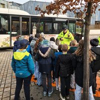 Eine Gruppen von Kindern hört einem Busfahrer zu, der vor einem Bus spricht. Der Bus steht an einer Haltestelle, während ein Baum im Hintergrund sichtbar ist. Die Kinder sind in bunten Jacken gekleidet und scheinen aufmerksam zu zuhören.