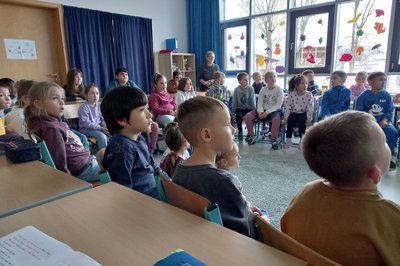 Eine Gruppe von Kindern sitzt in einem Klassenraum und schaut aufmerksam in eine Richtung. Die Atmosphäre wirkt konzentriert und interessiert. Die Wände sind mit bunten Bildern und Dekorationen geschmückt, während einige Kinder an Tischen sitzen und andere auf Stühlen stehen.