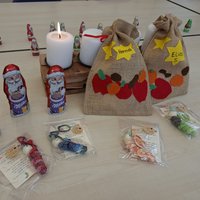Auf dem Tisch sind mehrere Schokoladenweihnachtsmänner, zwei leuchtende Kerzen und Geschenktüten aus Jute mit Namen darauf. Vor den Tüten liegen handgemachte Geschenke in durchsichtigen Tüten, koloriert in verschiedenen Farben.