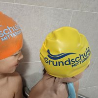 Zwei Kinder in Badekappen, eine orange und eine gelb, mit dem Aufdruck „Grundschule Mitterdorf“. Sie schauen sich an, während sie sich in einem schwimmbadähnlichen Umfeld befinden.