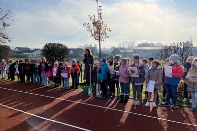 Eine Gruppe von Kindern steht auf einer Laufbahn im Freien. Sie sind in warmen Winterkleidung gehüllt und schauen aufmerksam in die Richtung einer Lehrerin, die neben einem kleinen Baum steht. Die Szenerie vermittelt eine festliche Atmosphäre, möglicherweise zu einem speziellen Anlass wie einer Baumpflanzung.