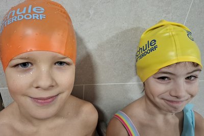 Zwei Kinder stehen nebeneinander an einer Wand. Das erste Kind trägt eine orangefarbene Badekappe mit der Aufschrift "Schule Hitterdorf", das zweite eine gelbe Badekappe. Beide lächeln freundlich in die Kamera, während sie in Freizeitkleidung abgebildet sind.