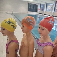 Vier Kinder in Badebekleidung stehen nebeneinander am Beckenrand. Jedes Kind trägt eine farbige Badekappe mit dem Aufdruck „Grundschule“. Im Hintergrund ist ein Schwimmbecken sichtbar. Die Kinder scheinen auf ihre Schwimmstunde zu warten.