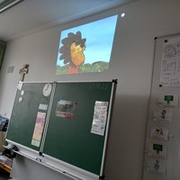 In einem Klassenzimmer wird ein Projektor verwendet, um eine Animation eines Löwen an die Wand zu projizieren. Der Löwe hat eine auffällige Mähne und schaut nachdenklich in die Ferne. Im Vordergrund steht eine Tafel mit weiteren Unterrichtsmaterialien.