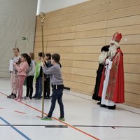 Eine Gruppe von Kindern steht in einer Turnhalle und präsentiert etwas. Rechts steht eine Person in einem festlichen Kleid mit Mitra und Bischofsstab, die traditionell den Nikolaus darstellt. Im Hintergrund sind weitere Kinder zu sehen, die auf dem Boden sitzen.