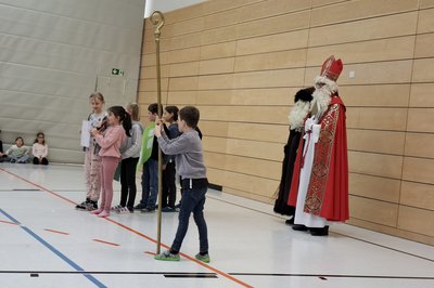 Eine Gruppe von Kindern steht in einer Turnhalle und präsentiert etwas. Rechts steht eine Person in einem festlichen Kleid mit Mitra und Bischofsstab, die traditionell den Nikolaus darstellt. Im Hintergrund sind weitere Kinder zu sehen, die auf dem Boden sitzen.