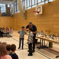 Eine Gruppe von Kindern sitzt auf dem Boden einer Sporthalle und beobachtet zwei Kinder, die vor einem Erwachsenen stehen. Der Erwachsene erklärt etwas und zeigt auf eine Reihe von Figuren auf einem Tisch. Die Atmosphäre ist konzentriert und interessert.