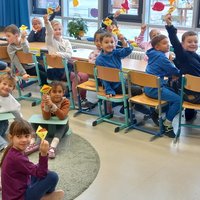 In einem Klassenraum sitzen Kinder an Tischen und halten Karten in die Höhe. Die Mehrheit blickt lächelnd zur Kamera. Die Umgebung wirkt freundlich und einladend, mit viel natürlichem Licht, das durch die Fenster strömt.