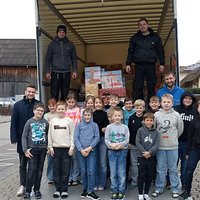Eine Gruppe von Kindern und Erwachsenen steht vor einem Lkw mit Kisten und Geschenken im Inneren. Die Kinder lächeln und posieren fröhlich, während zwei Erwachsene im Lkw stehen. Im Hintergrund sind Gebäude und eine Straße sichtbar.