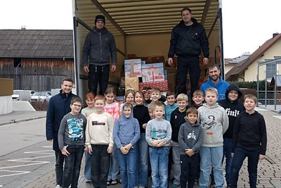 Eine Gruppe von Kindern und Erwachsenen steht vor einem Lkw mit Kisten und Geschenken im Inneren. Die Kinder lächeln und posieren fröhlich, während zwei Erwachsene im Lkw stehen. Im Hintergrund sind Gebäude und eine Straße sichtbar.