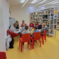 Eine Gruppe von Kindern sitzt auf roten Stühlen in einer Bibliothek. Sie sind in eine Diskussion vertieft, während Bücherregale im Hintergrund sichtbar sind. Die Atmosphäre ist freundlich und einladend, ideal für gemeinsames Lernen und Austausch.