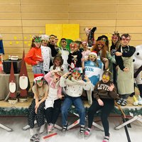 Eine Gruppe von 16 Personen steht auf einer Bühne, die festlich dekoriert ist. Sie tragen bunte Masken und verschiedene Kostüme. Einige haben Hüte auf, während sie fröhlich posieren. Im Hintergrund sind festliche Elemente wie Kerzen und eine weihnachtliche Dekoration sichtbar.