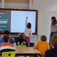 Ein Kind zeigt mit dem Finger auf ein Wortsuchrätsel an einer interaktiven Tafel in einem Klassenzimmer. Im Hintergrund stehen mehrere Schüler, während eine Lehrerin daneben steht. Das Thema des Rätsels sind Weihnachtsbegriffe.