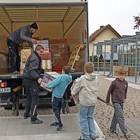 Fünf Personen, darunter Kinder und Erwachsene, stehen vor einem Lkw. Sie laden Geschenke aus dem Lkw auf die Rampe. Die Umgebung wirkt ruhig und freundlich, mit Wohngebäuden im Hintergrund.
