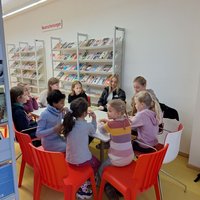 Eine Gruppe von Kindern und einem Erwachsenen sitzt zusammen an einem Tisch in einem Raum, umgeben von Bücherregalen. Sie scheinen an einer Aktivität teilzunehmen, die gemeinschaftliches Lernen oder Spielen fördert. Die Atmosphäre wirkt freundlich und einladend.