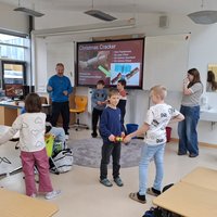 Eine Gruppe von Kindern steht in einem Klassenraum und zieht an Weihnachtsknallern. Im Hintergrund ist eine Präsentation zu sehen, die Informationen über Weihnachtsknaller zeigt. Ein Erwachsener steht in der Nähe und beobachtet die Aktivitäten der Kinder.