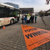 Eine Gruppe von Schülern steht in der Nähe eines Stadtbusses, während ein Verkehrssicherheitstrainer ihnen etwas erklärt. Vor dem Bus liegt ein orangefarbenes Banner mit der Aufschrift „Toter Winkel“. Die Szene spielt sich in einer städtischen Umgebung ab.