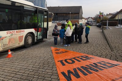 Eine Gruppe von Schülern steht in der Nähe eines Stadtbusses, während ein Verkehrssicherheitstrainer ihnen etwas erklärt. Vor dem Bus liegt ein orangefarbenes Banner mit der Aufschrift „Toter Winkel“. Die Szene spielt sich in einer städtischen Umgebung ab.