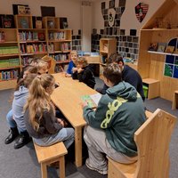 Eine Gruppe von sieben Kindern sitzt an einem langen Holztisch in einer Bibliothek. Sie sind in ein Gespräch vertieft und betrachten ein Buch. Im Hintergrund sind Regale mit vielen Büchern und dekorativen Elementen in der Wand zu sehen.