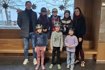 Eine Gruppe von Kindern und zwei Erwachsenen steht in einem Hausflur vor einem Fenster. Alle tragen Helme. Die Kinder lächeln und wirken fröhlich. Im Hintergrund sind Fenster mit Blick auf Bäume und winterliche Dekoration.