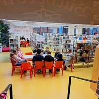 In einer Bibliothek sind mehrere Kinder und Jugendliche um einen Tisch mit roten Stühlen versammelt. Im Hintergrund sind Bücherregale sichtbar. Die Atmosphäre ist freundlich und einladend, ideal für gemeinsames Lernen oder Diskussionen.