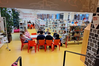 In einer Bibliothek sind mehrere Kinder und Jugendliche um einen Tisch mit roten Stühlen versammelt. Im Hintergrund sind Bücherregale sichtbar. Die Atmosphäre ist freundlich und einladend, ideal für gemeinsames Lernen oder Diskussionen.