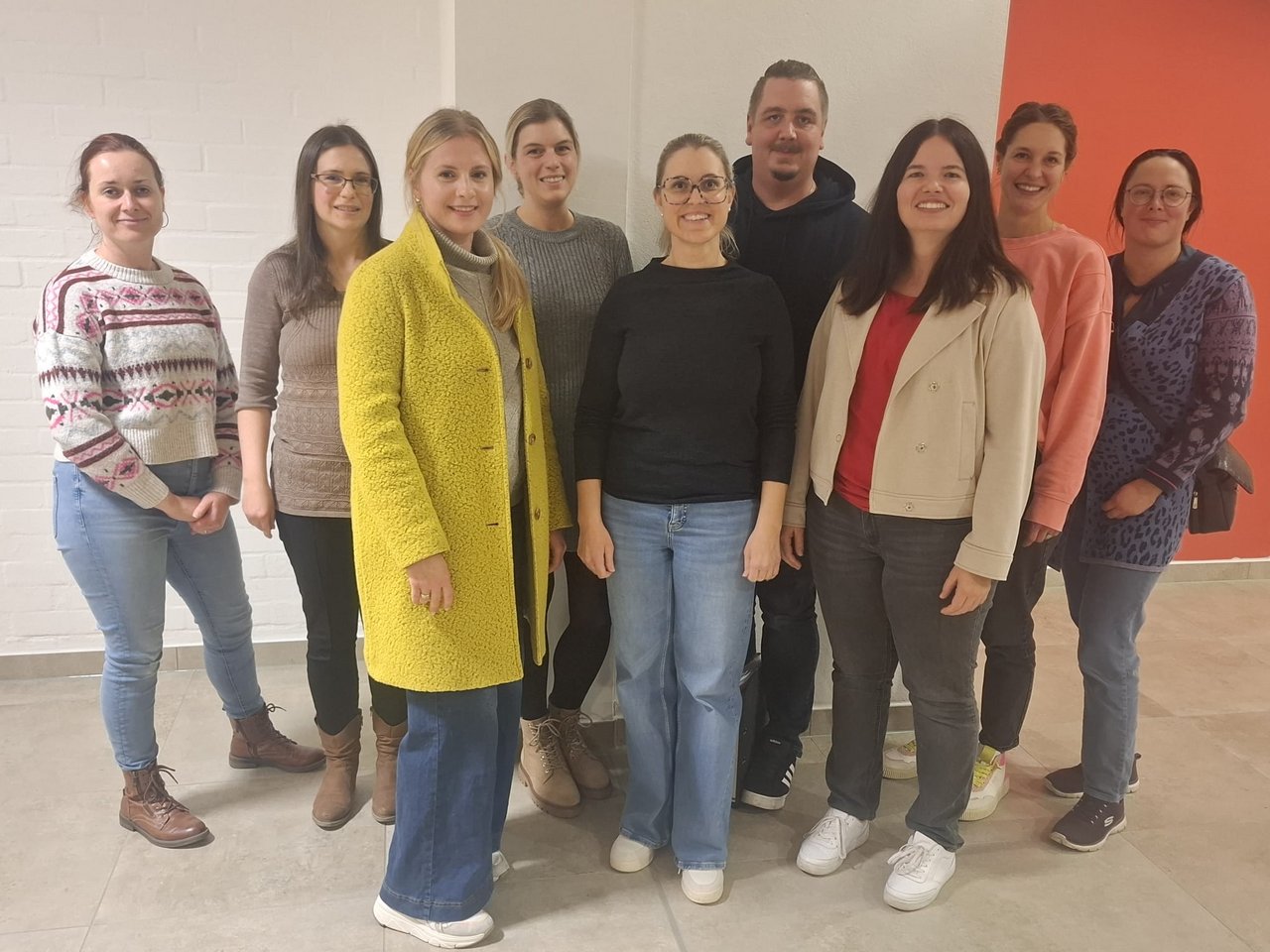 Eine Gruppe von zehn Personen steht in einem Raum. Die Frauen sind unterschiedlich gekleidet, einige tragen Pullover oder Jacken, andere sind legere. Im Hintergrund sind helle Wände und ein roter Bereich zu sehen. Alle lächeln in die Kamera.