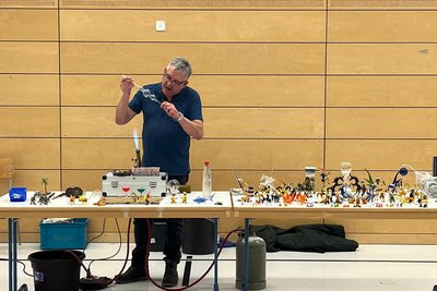 Ein Dozent steht an einem Tisch und demonstriert eine experimentelle Technik, während er ein Glasobjekt hält. Auf dem Tisch sind verschiedene bunte Figuren und Materialien verteilt, die für das Experiment verwendet werden. Im Hintergrund sind aufmerksame Kinder zu sehen.