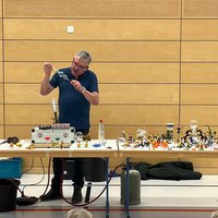 Ein Dozent steht an einem Tisch und demonstriert eine experimentelle Technik, während er ein Glasobjekt hält. Auf dem Tisch sind verschiedene bunte Figuren und Materialien verteilt, die für das Experiment verwendet werden. Im Hintergrund sind aufmerksame Kinder zu sehen.