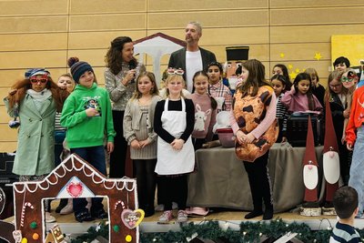 Eine Gruppe von Kindern und Erwachsenen steht auf einer Bühne, umgeben von weihnachtlicher Dekoration. Die Kinder tragen verschiedene Kostüme und halten Requisiten. Im Hintergrund sind einige musikalische Instrumente und eine Dekoration zu sehen, die an ein Lebkuchenhaus erinnert.