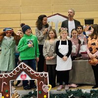 Eine Gruppe von Kindern und Erwachsenen steht auf einer Bühne, umgeben von weihnachtlicher Dekoration. Die Kinder tragen verschiedene Kostüme und halten Requisiten. Im Hintergrund sind einige musikalische Instrumente und eine Dekoration zu sehen, die an ein Lebkuchenhaus erinnert.