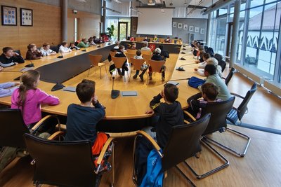 Eine Gruppe von Kindern sitzt in einem großen Konferenzraum an einem langen Tisch. Sie schauen zur Vorderseite des Raumes, wo eine Präsentation vorbereitet ist. Der Raum hat große Fenster, die natürliches Licht hereinlassen.