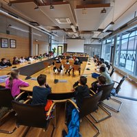 Eine Gruppe von Kindern sitzt in einem großen Konferenzraum an einem langen Tisch. Sie schauen zur Vorderseite des Raumes, wo eine Präsentation vorbereitet ist. Der Raum hat große Fenster, die natürliches Licht hereinlassen.