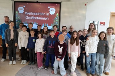 Eine Gruppe von Kindern und Erwachsenen steht lächelnd vor einer Präsentation mit dem Titel "Weihnachten in Großbritannien". Die Kinder tragen unterschiedliche Kleidung und sind in einem hellen Raum. Im Hintergrund sind weihnachtliche Dekorationen sichtbar.