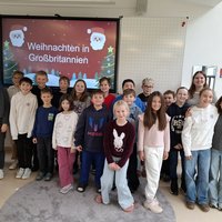 Eine Gruppe von Kindern und Erwachsenen steht lächelnd vor einer Präsentation mit dem Titel "Weihnachten in Großbritannien". Die Kinder tragen unterschiedliche Kleidung und sind in einem hellen Raum. Im Hintergrund sind weihnachtliche Dekorationen sichtbar.