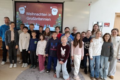 Eine Gruppe von Kindern und Erwachsenen steht lächelnd vor einer Präsentation mit dem Titel "Weihnachten in Großbritannien". Die Kinder tragen unterschiedliche Kleidung und sind in einem hellen Raum. Im Hintergrund sind weihnachtliche Dekorationen sichtbar.