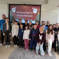 Eine Gruppe von Kindern und Erwachsenen steht lächelnd vor einer Präsentation mit dem Titel "Weihnachten in Großbritannien". Die Kinder tragen unterschiedliche Kleidung und sind in einem hellen Raum. Im Hintergrund sind weihnachtliche Dekorationen sichtbar.