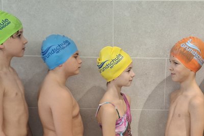 Eine Gruppe von sieben Kindern steht in einer Reihe in Badesachen und Schwimmkappen. Sie blicken einander an. Die Kappen sind in verschiedenen Farben: grün, blau, gelb und orange. Die Kinder stehen in einem hellen Raum mit einem fliesenartigen Hintergrund.